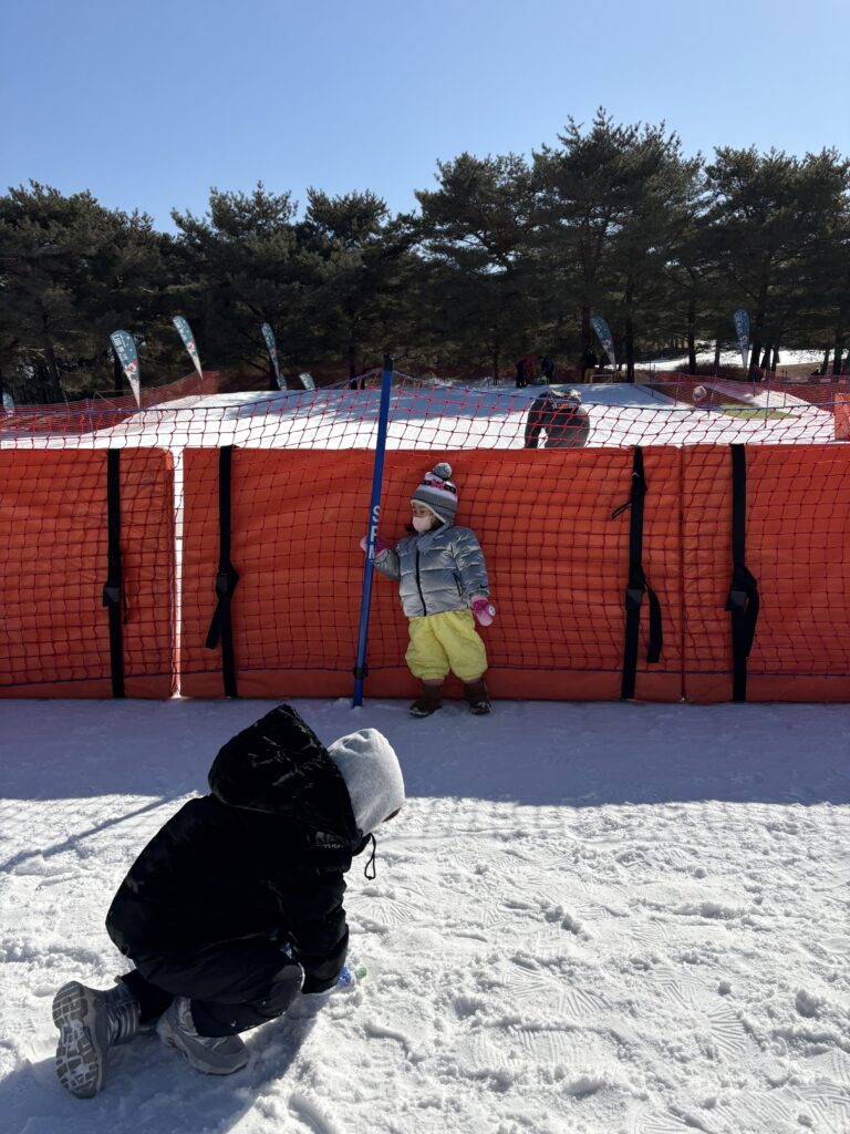 눈 위에서 놀고 있는 아이들. 한 아이는 빨간 울타리에 기대고 있고, 다른 아이는 눈을 만지고 있다.